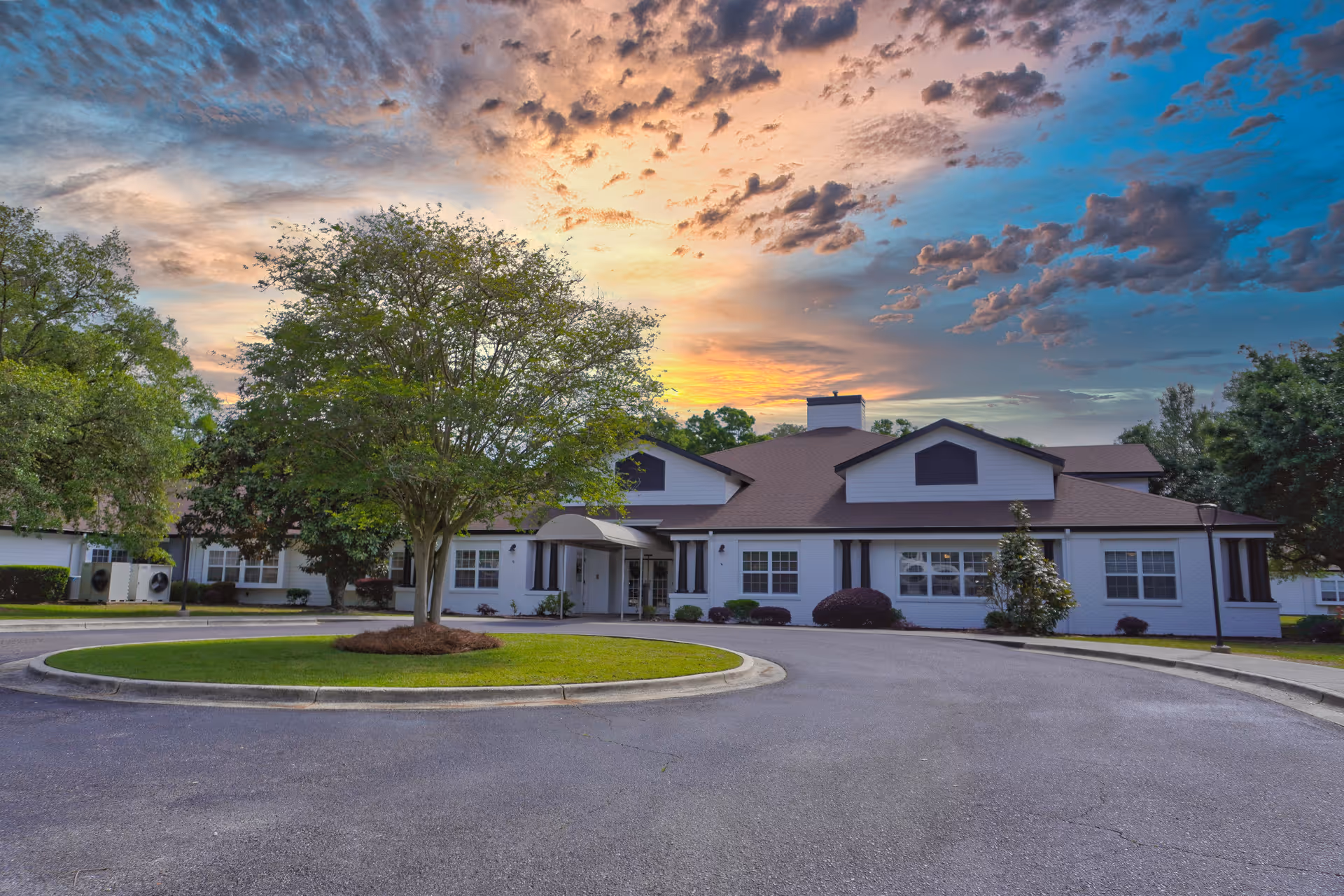 Exterior view of a single-story senior living facility building with a circular driveway and a tree in the center, under a colorful sunset sky with scattered clouds.