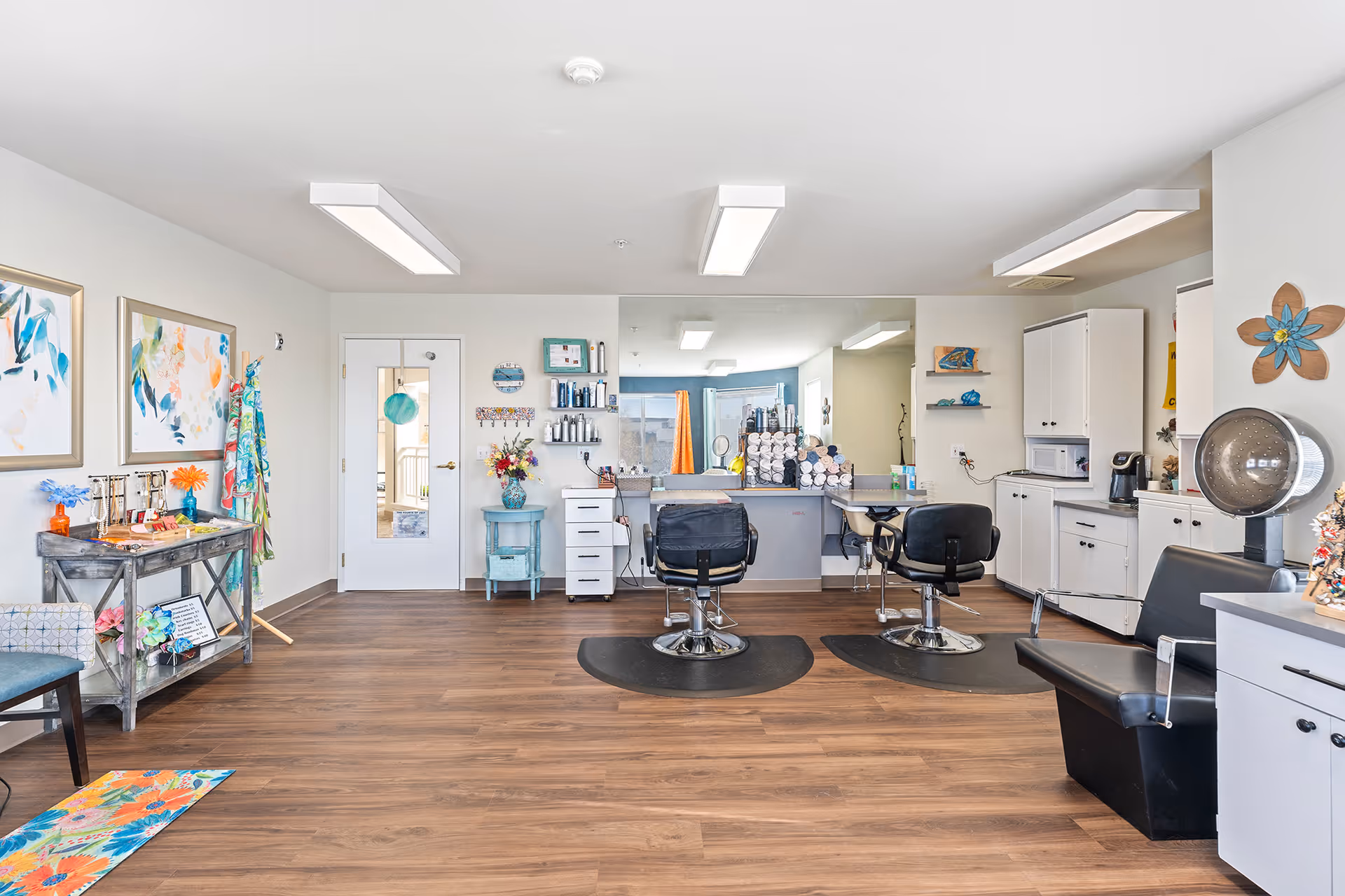 A bright and clean salon area inside a senior living facility featuring two black salon chairs in front of a large mirror, a hair dryer chair, white cabinets, and shelves with hair care products and towels. The room has wooden flooring, colorful artwork on the walls, and a small table with decorative items and flowers.