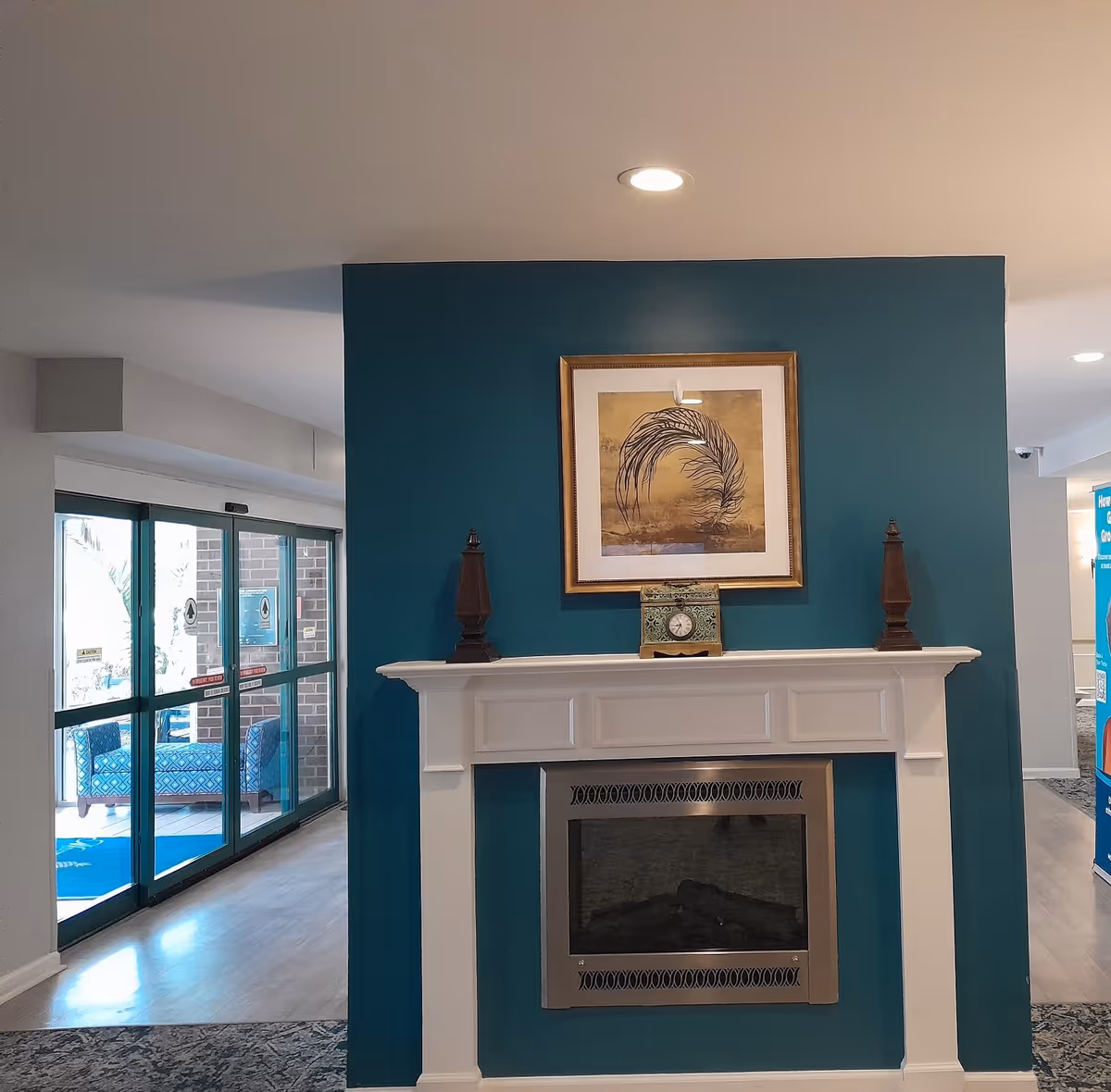Interior view of a senior living facility lobby area featuring a white fireplace with a teal accent wall behind it. On the mantel are two decorative wooden finials and a small clock. Above the mantel hangs a framed artwork depicting a feather. To the left, there are glass sliding doors leading outside, and to the right, part of a hallway and a blue informational sign are visible.
