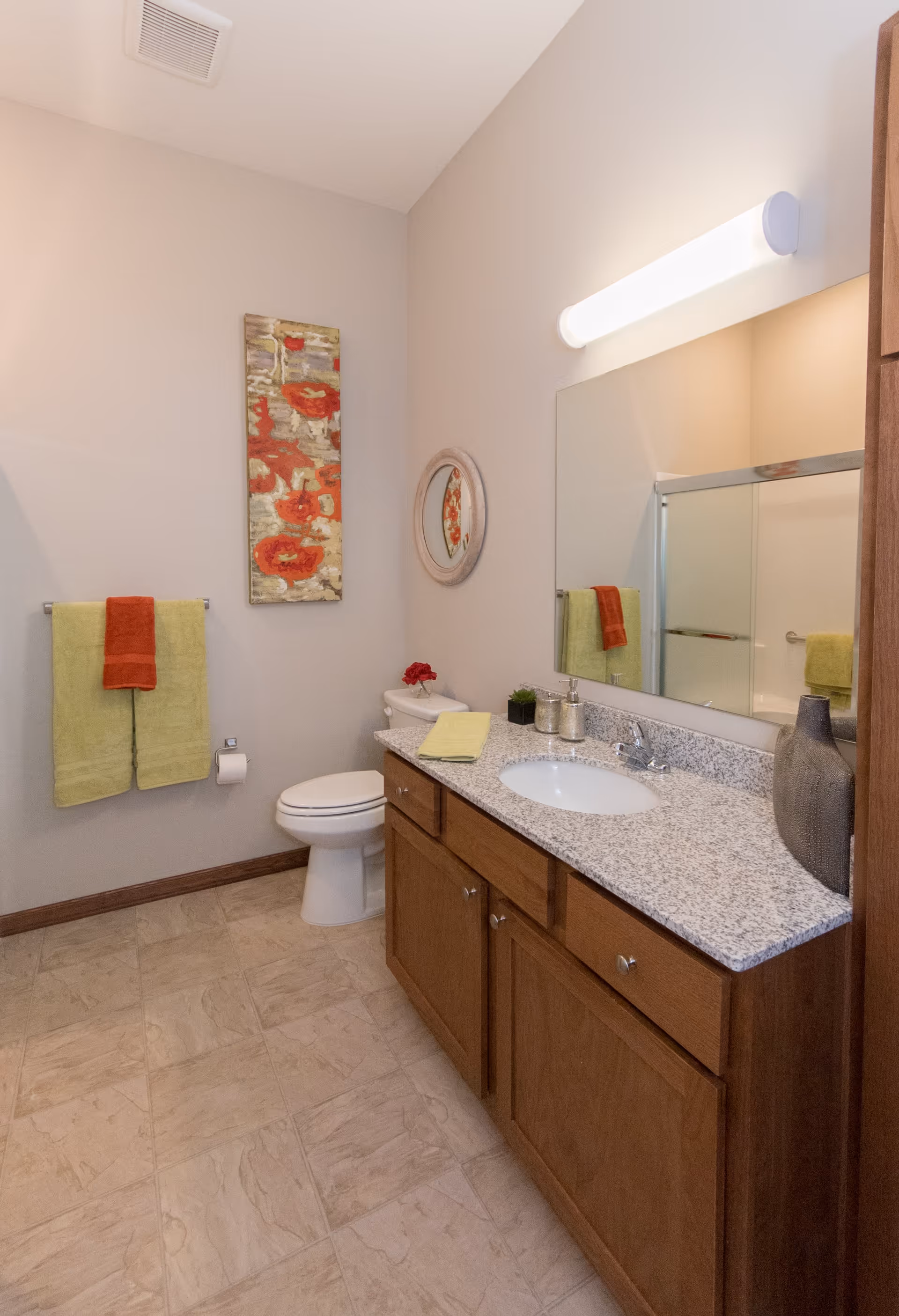 A clean bathroom with a granite countertop vanity featuring a sink, soap dispenser, and decorative vase. There is a toilet next to the vanity, a towel rack with green and red towels, and wall art with red flowers. The bathroom has beige walls and tiled flooring.