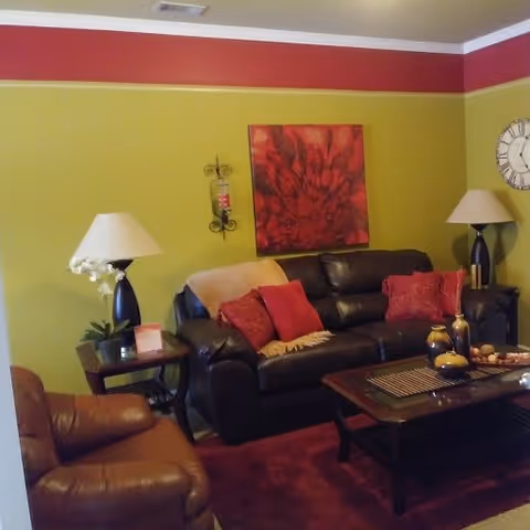 Cozy living room with a dark leather sofa and armchair, red accent pillows, coffee table, lamps, and red floral wall art.