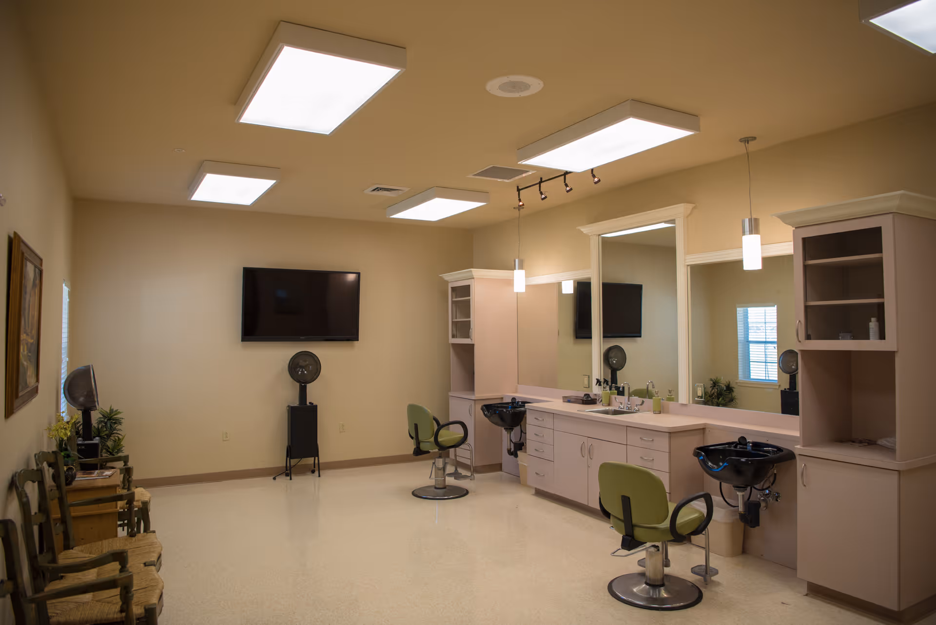 Interior of a salon area in a healthcare facility with two green salon chairs in front of black hair washing sinks, large mirrors, cabinets, a wall-mounted TV, and several ceiling lights.