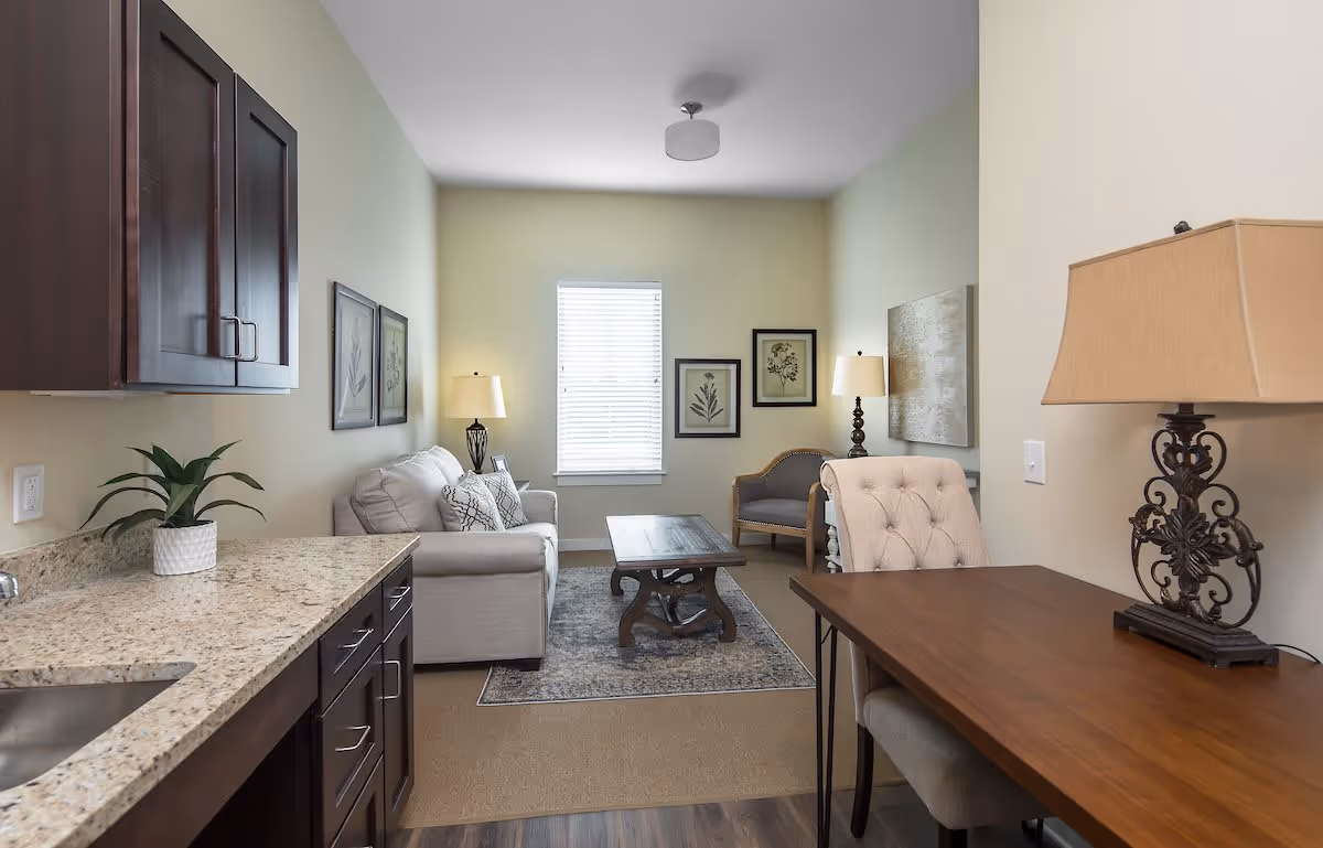 Open living area with a sofa, coffee table, armchairs, desk, and a kitchenette with a granite countertop.