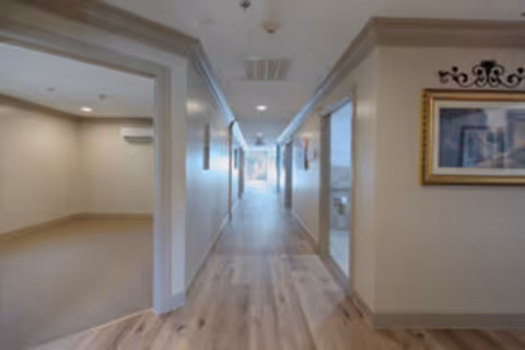 Long interior hallway in a senior living facility with wood flooring, open doorways, and framed artwork on the wall.