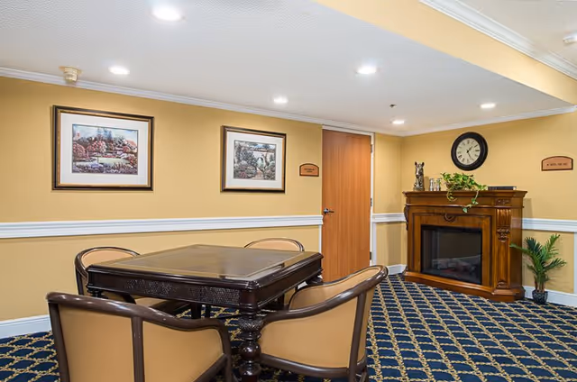 A cozy interior room with a dark wooden table surrounded by four beige and brown chairs on a patterned carpet. The walls are painted yellow with white trim and decorated with two framed landscape paintings. A wooden electric fireplace with decorative items and a clock above it is positioned against one wall, with a potted plant nearby.