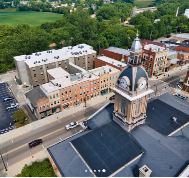 Aerial view of a small town with a prominent clock tower in the foreground, surrounded by historic brick buildings, a street with parked cars, and green trees in the background.