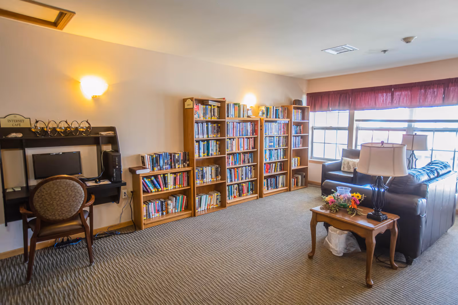 A cozy senior living common area featuring a small internet cafe with a computer and chair on the left, several wooden bookshelves filled with books along the wall, and a seating area with a dark leather couch and armchair near large windows with red curtains. A wooden side table with a lamp and flower arrangement is placed next to the couch.