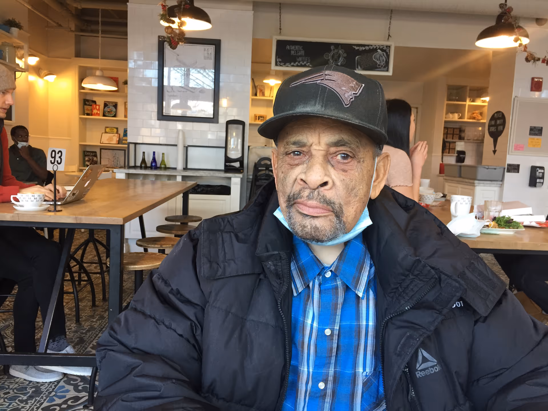 An elderly man wearing a black jacket, blue plaid shirt, and a dark baseball cap sits at a table in a cafe or dining area. Behind him, there are other people seated at tables, shelves with books and decorative items, and warm lighting fixtures.