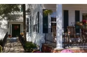 Front porch and entrance of a white house with columns, green shutters, potted plants, and outdoor seating.