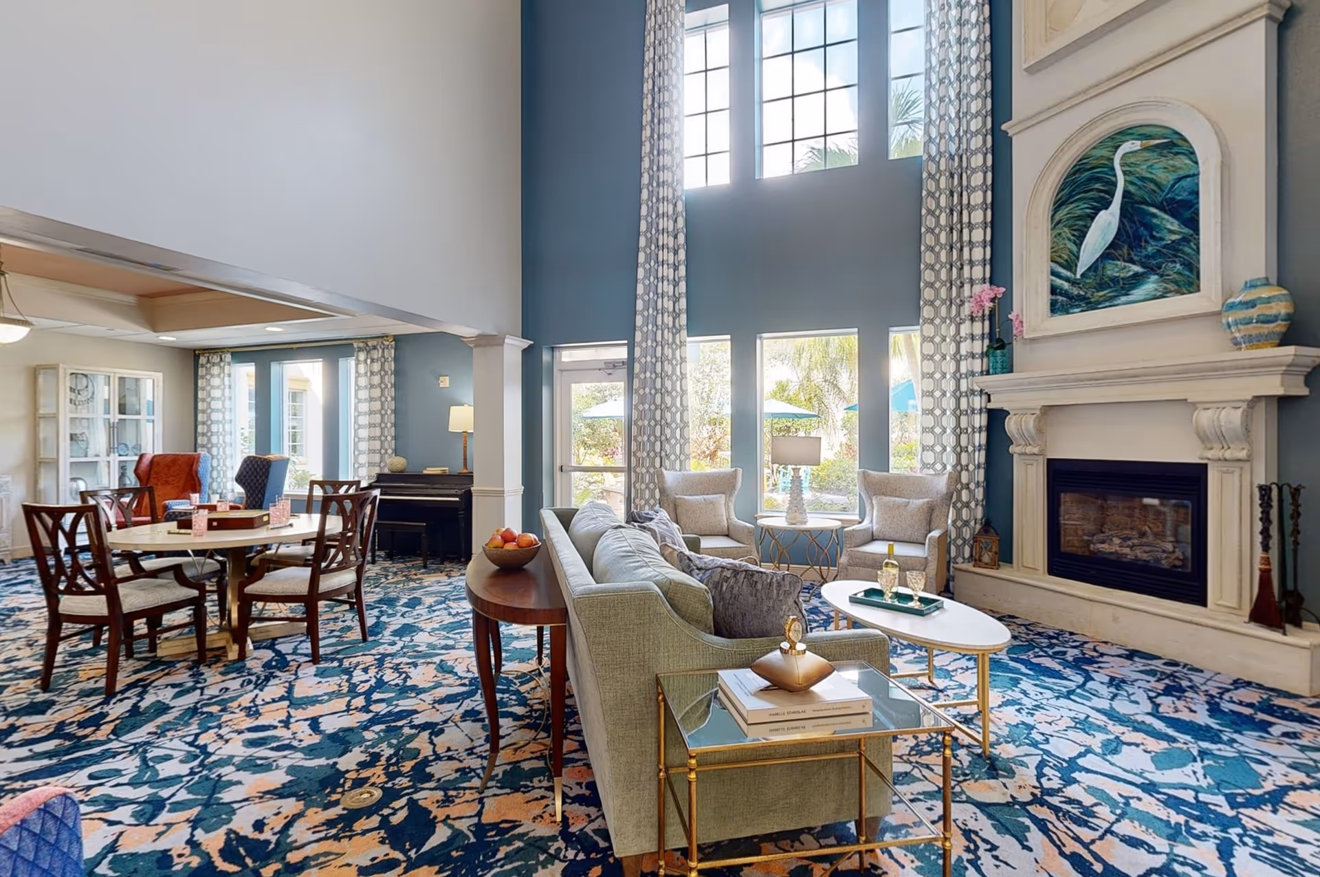 A spacious and well-lit living room area with high ceilings and large windows. The room features a light green sofa, two beige armchairs, a white oval coffee table, and a fireplace with artwork above it. There is a round dining table with chairs in the background, patterned carpet flooring, and tall curtains framing the windows.