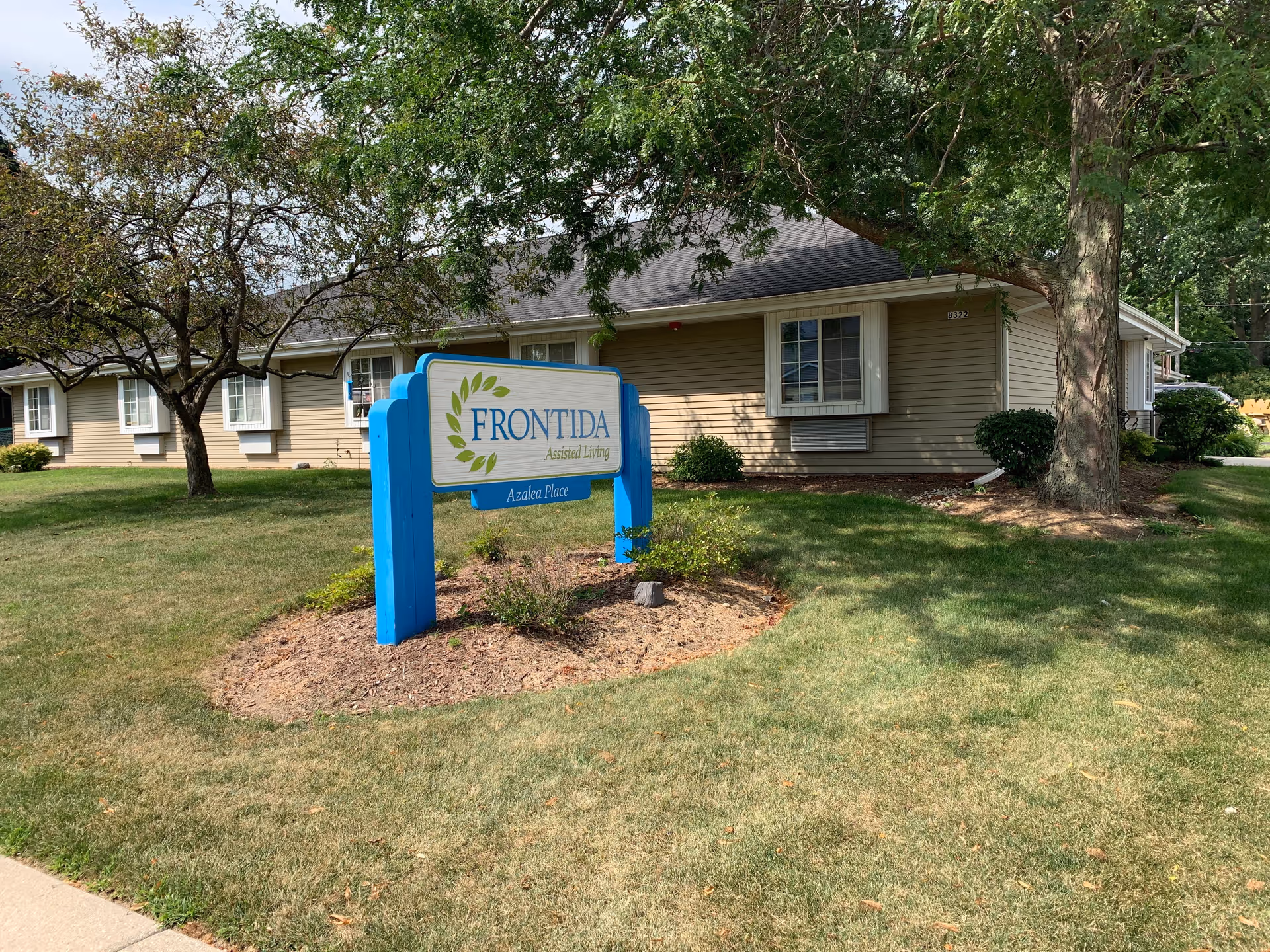 Exterior view of Frontida Assisted Living: Azalea Place building with a blue and white sign in front on a grassy lawn, surrounded by trees and bushes.