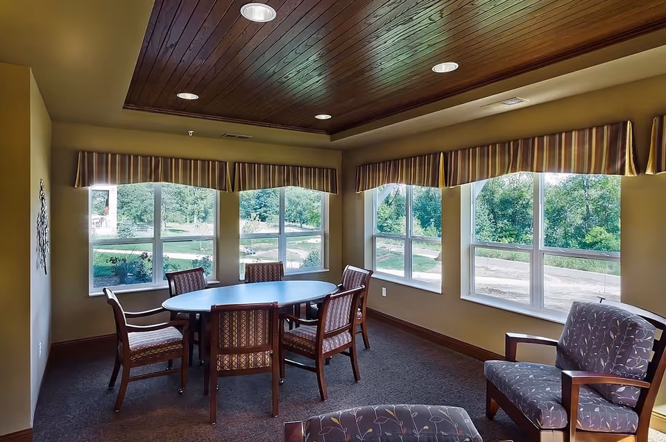 Communal sitting and dining area with an oval table surrounded by chairs and large windows looking out to greenery.