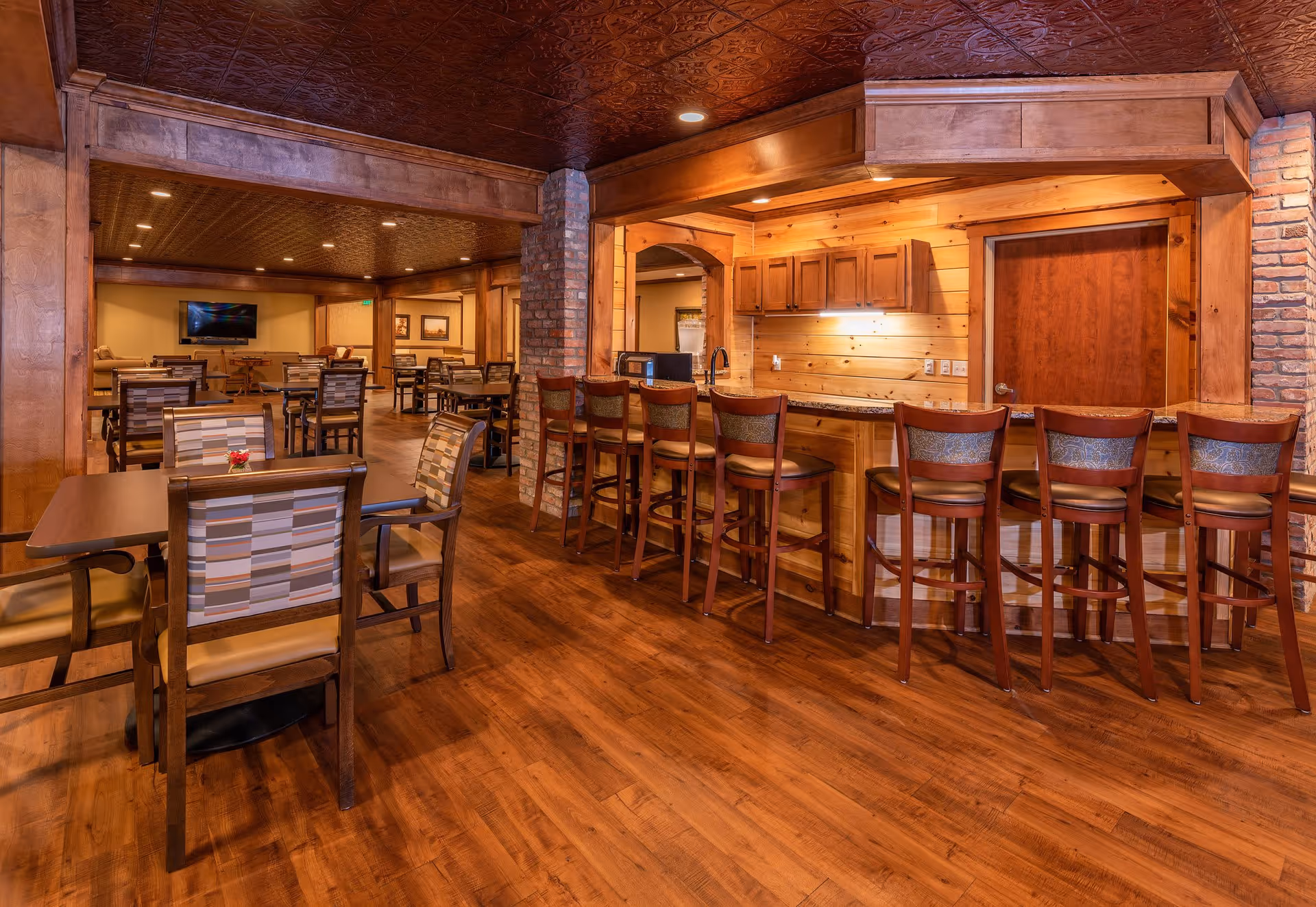 Interior view of a warm, wood-themed dining and lounge area with multiple tables and chairs, a bar counter with high chairs, and a TV mounted on the far wall.