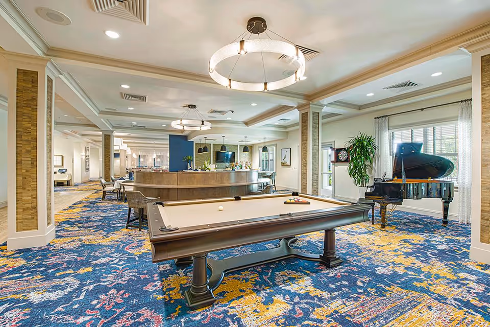 A spacious and well-lit recreational room featuring a pool table in the center, a grand piano near a window with sheer curtains, a dartboard on the wall, and a curved bar area with stools. The room has colorful patterned carpet, modern circular ceiling lights, and large windows allowing natural light.