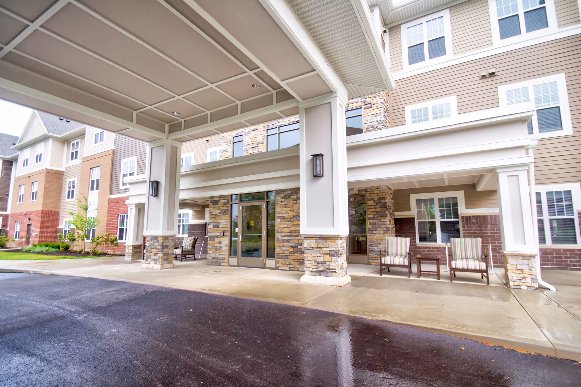 Covered entrance of a multi-story senior living building with stone pillars, glass doors, and outdoor seating.