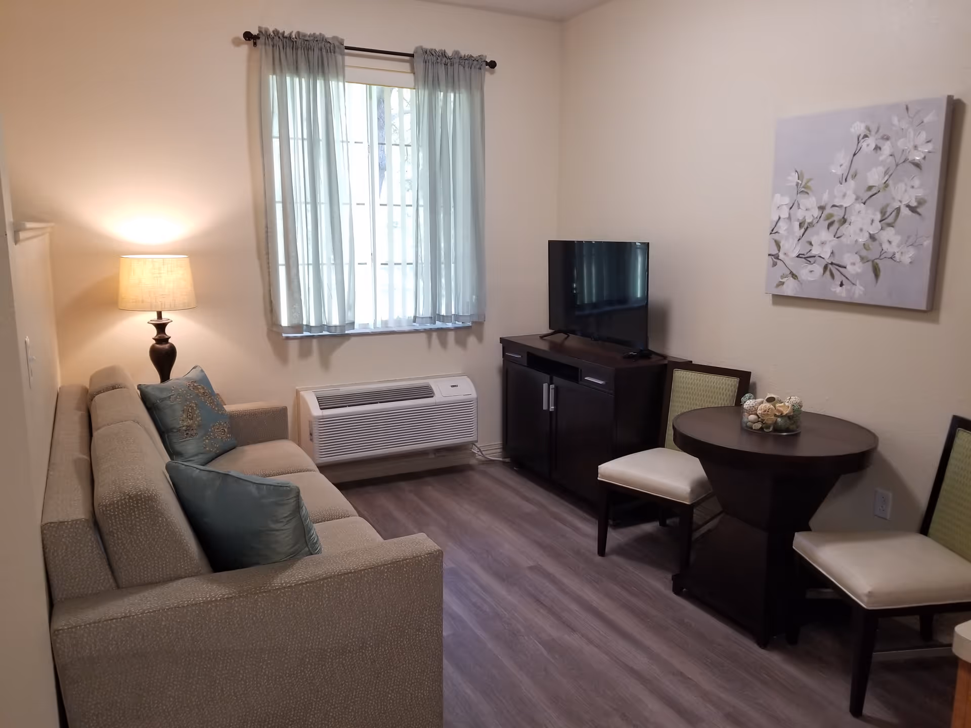 A small living room with a beige couch adorned with two blue-green pillows, a floor lamp beside the couch, a window with sheer curtains, a dark wood TV stand with a flat-screen TV, and a round dark wood table with two chairs. A floral painting hangs on the wall above the table.