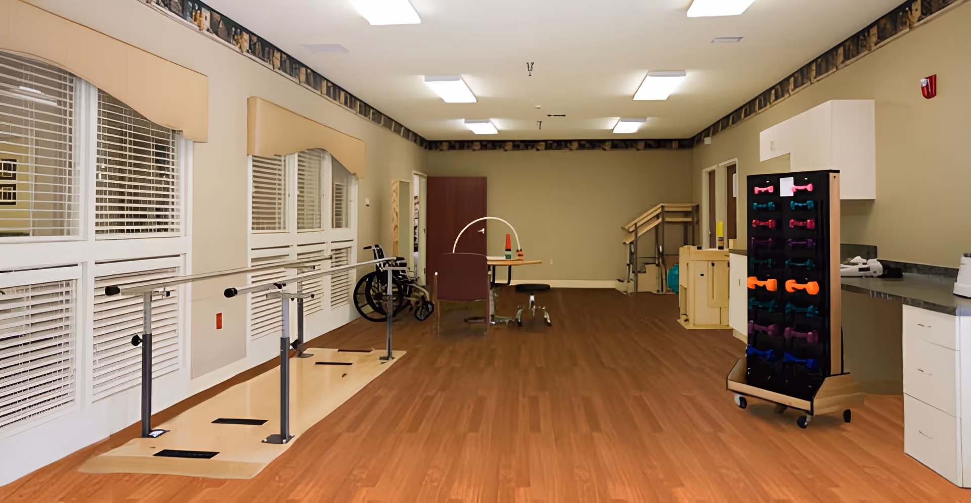 A rehabilitation room with wooden flooring, parallel bars for walking exercises along the left side near large windows with blinds. There is a wheelchair, a small table with chairs, and various exercise equipment including colorful hand weights on a rack. The room has beige walls and ceiling lights.