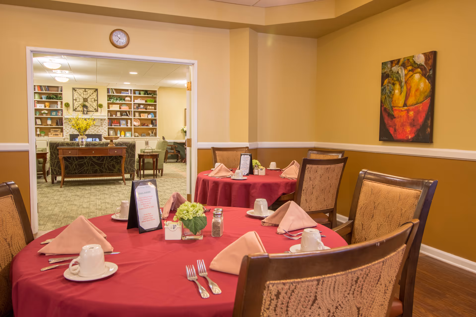 Dining room with round tables covered in red tablecloths, place settings, and chairs, opening into a lounge with bookshelves.