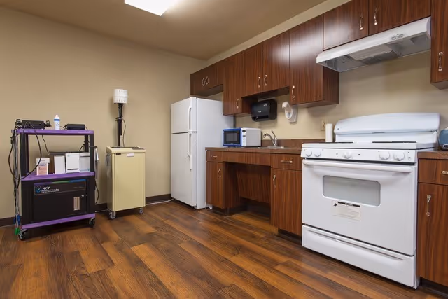 Interior view of a kitchen area with wooden cabinets, a white refrigerator, a white stove with an oven, a microwave on the countertop, and a small cart with medical or electronic equipment on the left side. The floor is wooden and the walls are beige.