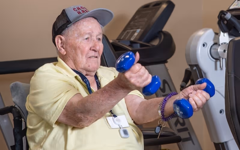 An elderly man wearing a yellow shirt and a gray cap is seated and lifting two small blue dumbbells in a fitness or rehabilitation room with exercise equipment in the background.