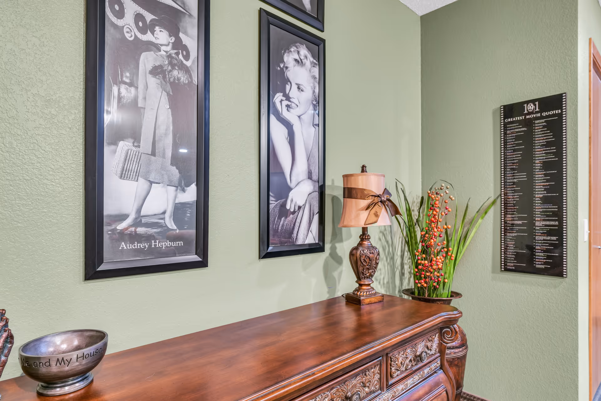 A corner of a room with light green walls featuring two black and white framed portraits of Audrey Hepburn and Marilyn Monroe. Below the portraits is an ornate wooden dresser with a decorative lamp and a potted plant with orange berries. On the right wall, there is a framed list titled '101 Greatest Movie Quotes.'