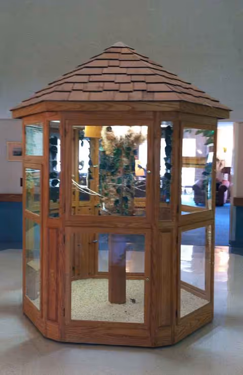 Wooden glass-paneled hexagonal display gazebo with a shingled roof inside a facility lobby.