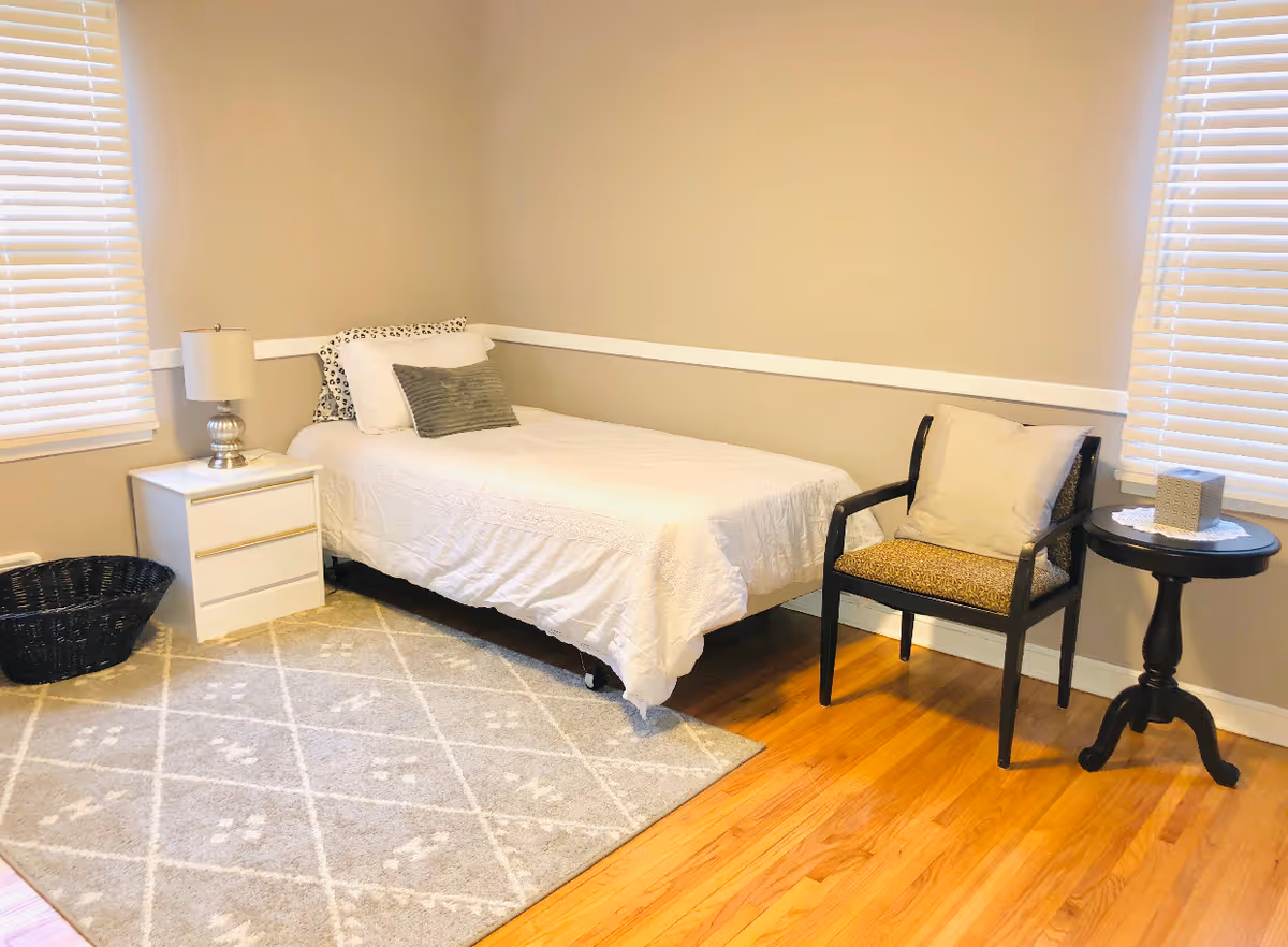 A cozy bedroom with a single bed covered in white bedding and pillows, a white nightstand with a silver lamp, a black woven basket, a patterned area rug, a wooden floor, a black chair with a cushion, and a small round black side table with a tissue box. The room has beige walls and two windows with white blinds.