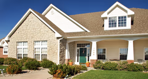 Exterior front view of a single-story residential building with a stone and siding facade, a green front door, multiple windows, and a well-maintained lawn and shrubs under a clear blue sky.