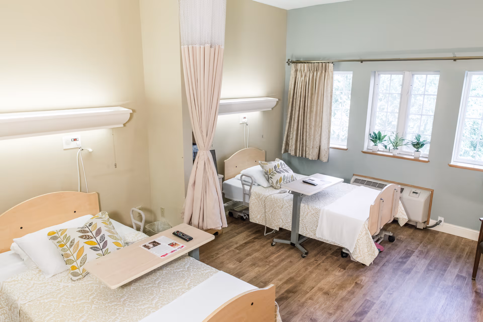 A bright nursing facility room with two hospital-style beds separated by a privacy curtain. Each bed has a patterned pillow and a beige bedspread. There are overbed tables with items like a remote control and papers. The room has wooden flooring, large windows with curtains, and small potted plants on the windowsill.