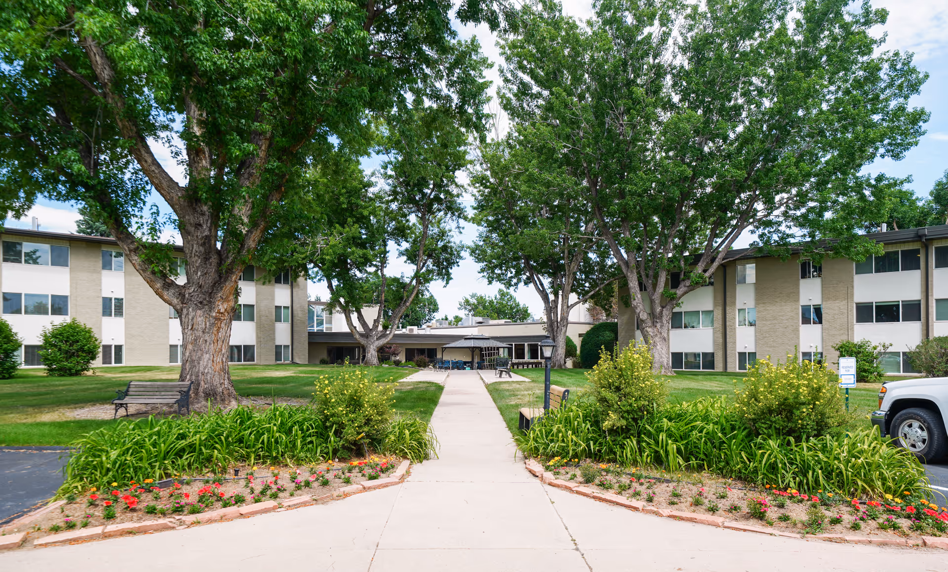 Exterior view of Winslow Court Assisted & Senior Living facility showing a three-story building with large windows, surrounded by green lawns, trees, flower beds, benches, and a paved walkway leading to the entrance.