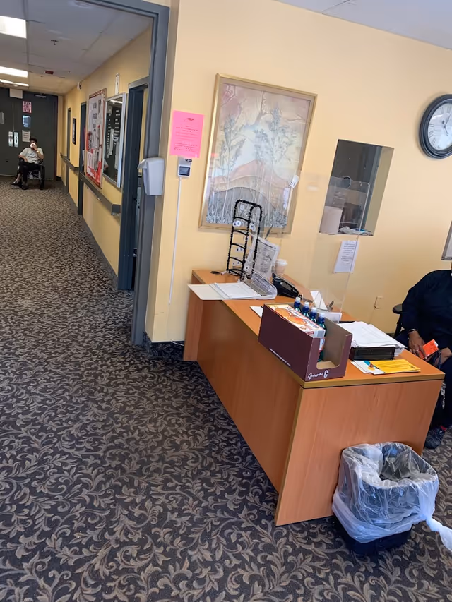 Interior view of a senior living facility hallway with patterned carpet and yellow walls. A wooden reception desk with papers, a box of snacks, and a clear protective barrier is on the right. A person is seated behind the desk, and another person in a wheelchair is visible further down the hallway. There is a framed artwork on the wall and a clock above the desk.