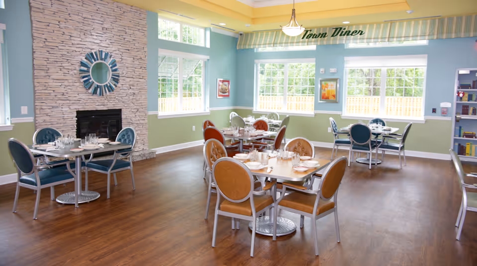 A bright dining room with several tables set for meals, featuring chairs with blue and brown upholstery. The room has large windows letting in natural light, a stone fireplace with a decorative round mirror above it, and a sign that reads 'Town Diner' on the wall. The walls are painted in light blue and green tones, and the floor is wooden.