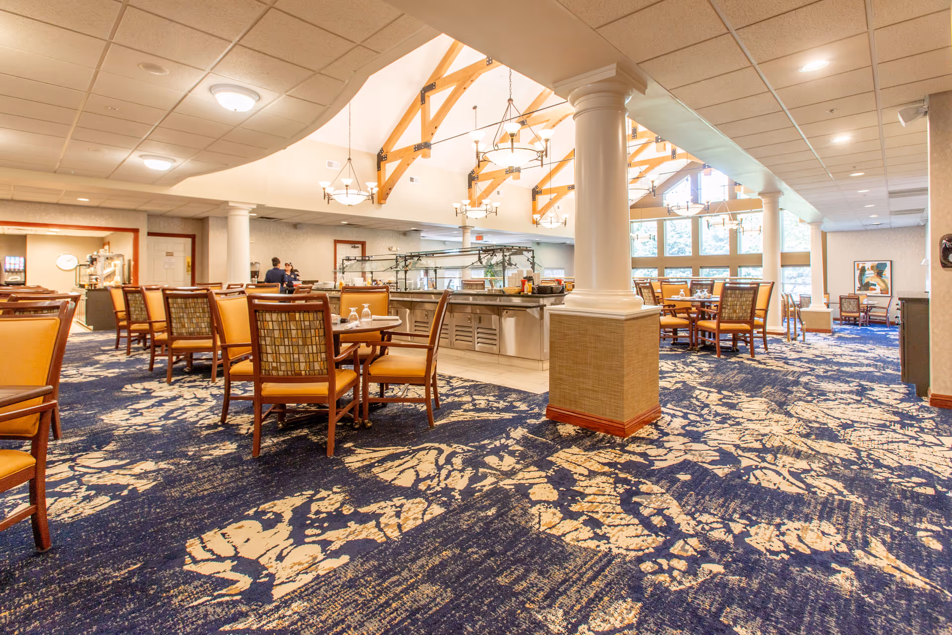 Spacious dining room with multiple tables and chairs arranged on a patterned blue and beige carpet. The room features high ceilings with wooden beams and several hanging light fixtures. Large windows allow natural light to fill the space. A buffet area with glass sneeze guards is visible in the background, along with two people near the serving area.