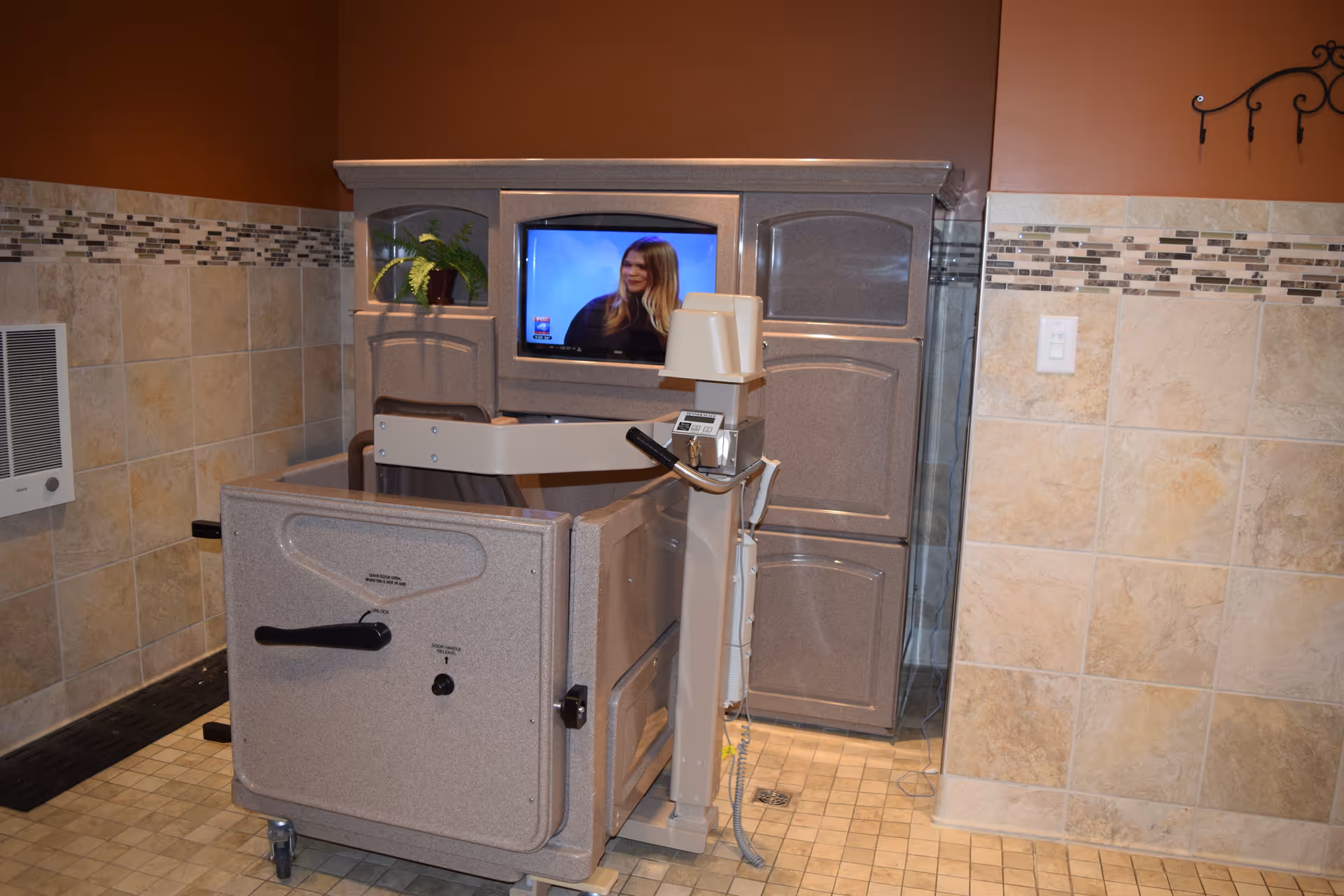 A bathing chair lift in a tiled bathroom with beige and brown walls. The chair is positioned in front of a cabinet with a TV screen showing a woman with long blonde hair. There is a small potted plant on the cabinet shelf and a wall-mounted heater on the left side.