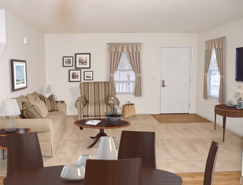 A cozy living room area with a beige sofa, a striped armchair, a wooden coffee table with a plant and magazine, and a round dining table with chairs in the foreground. The room has light-colored walls, two windows with beige curtains, framed pictures on the wall, and a white front door.