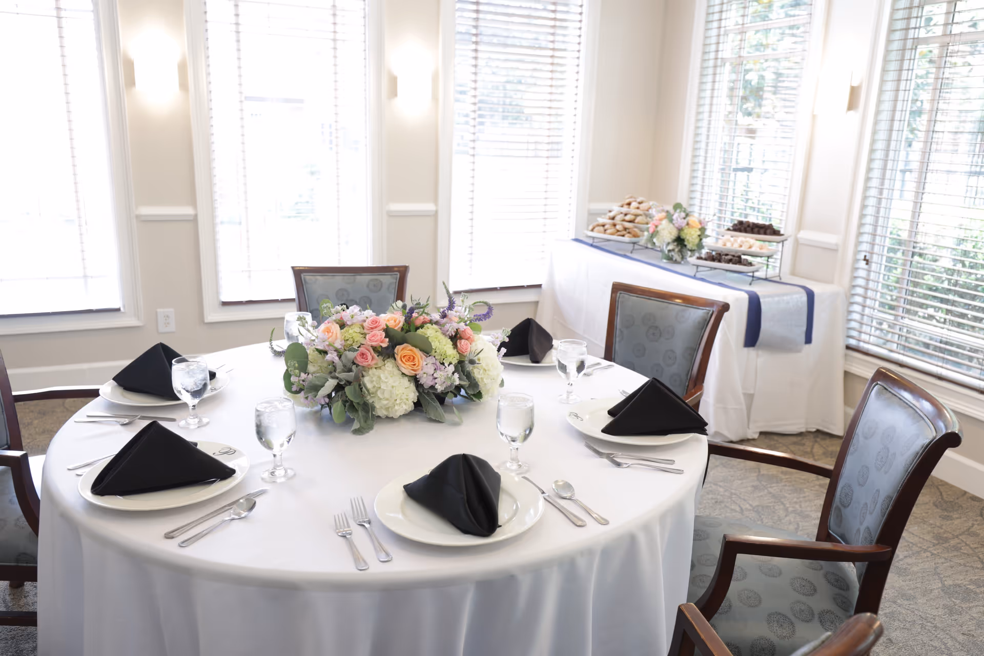 A round dining table set for five with white tablecloth, black folded napkins on white plates, silverware, and water glasses. A floral centerpiece with pink, peach, and white flowers decorates the middle of the table. In the background, a buffet table with trays of cookies and chocolates is visible near large windows with blinds.