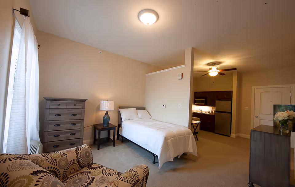 A senior living facility room with a single bed covered in white bedding, a wooden dresser, a side table with a lamp, a patterned armchair, and a small kitchenette area with a refrigerator, microwave, and cabinets. The room has beige walls and carpeted floor, with a ceiling light and a ceiling fan above the kitchenette.