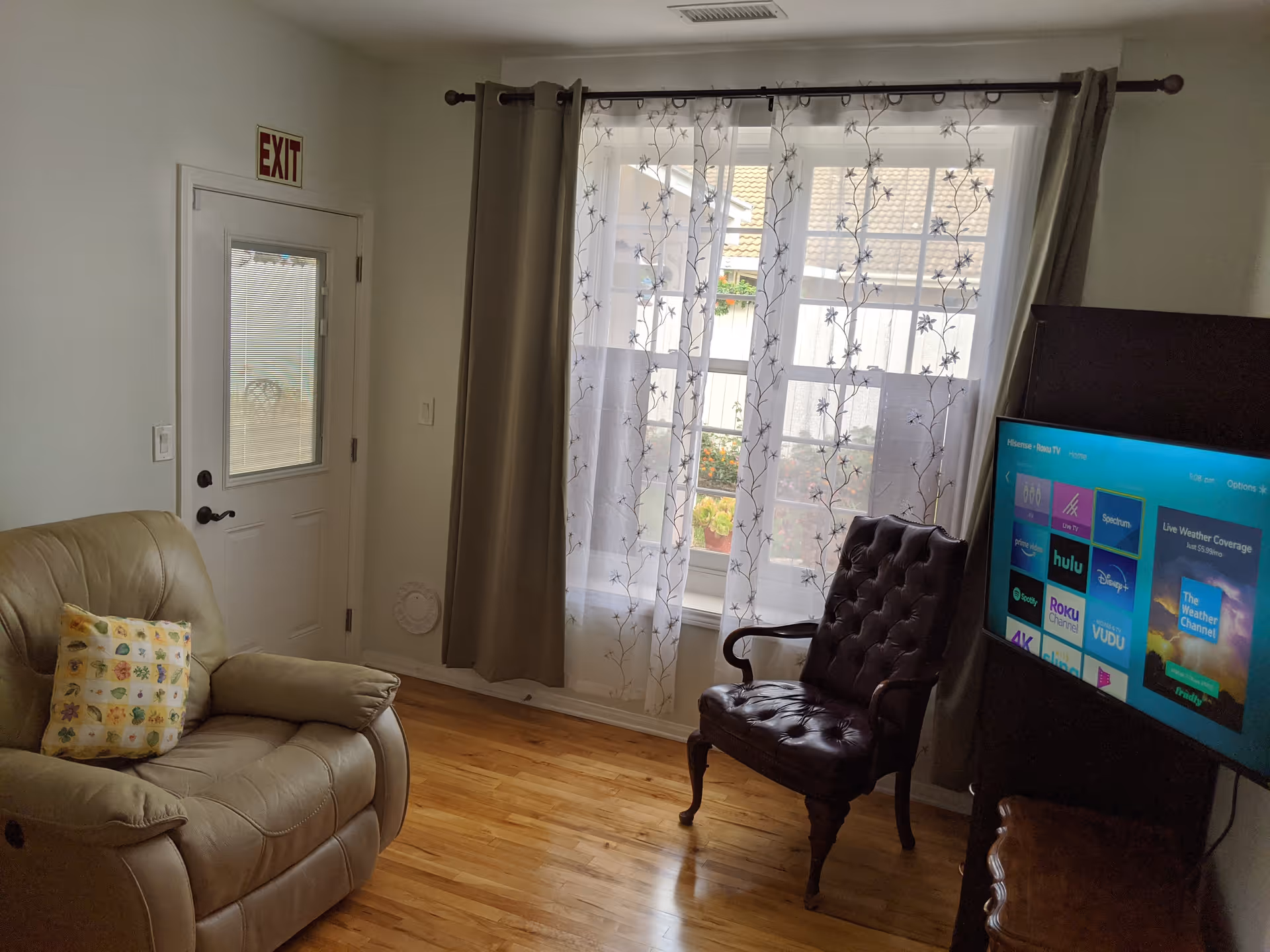 A cozy living room with a beige recliner chair with a floral pillow, a dark brown tufted armchair, a flat-screen TV displaying streaming apps, a window with sheer floral curtains and beige drapes, and a door with an exit sign above it.