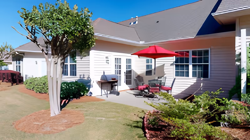 Outdoor patio of a single-story building with seating, a red umbrella, grill, and landscaped lawn.