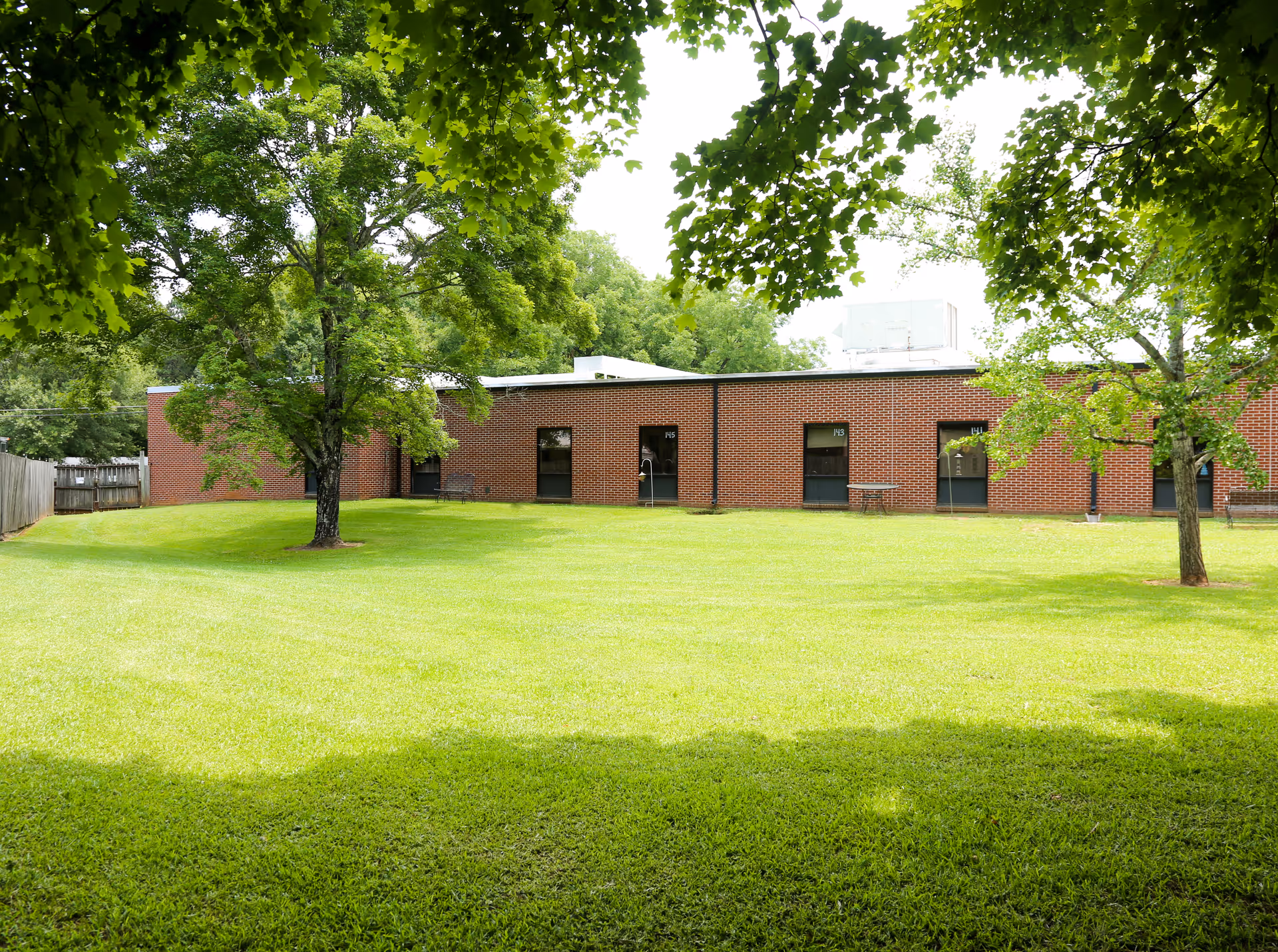 A grassy outdoor area with several large green trees providing shade. In the background, there is a single-story brick building with multiple doors and windows. The scene is bright and sunny, showing a peaceful and well-maintained lawn.