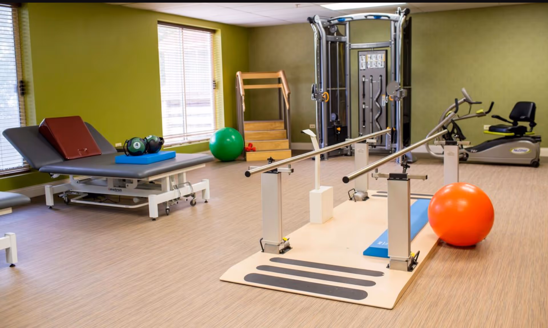 A well-lit physical therapy or exercise room with green walls and wooden flooring. The room contains parallel bars for walking exercises, a large orange exercise ball, a green exercise ball, a padded therapy table with weights on it, a recumbent exercise bike, and a multi-functional exercise machine. Two windows with blinds allow natural light into the room.