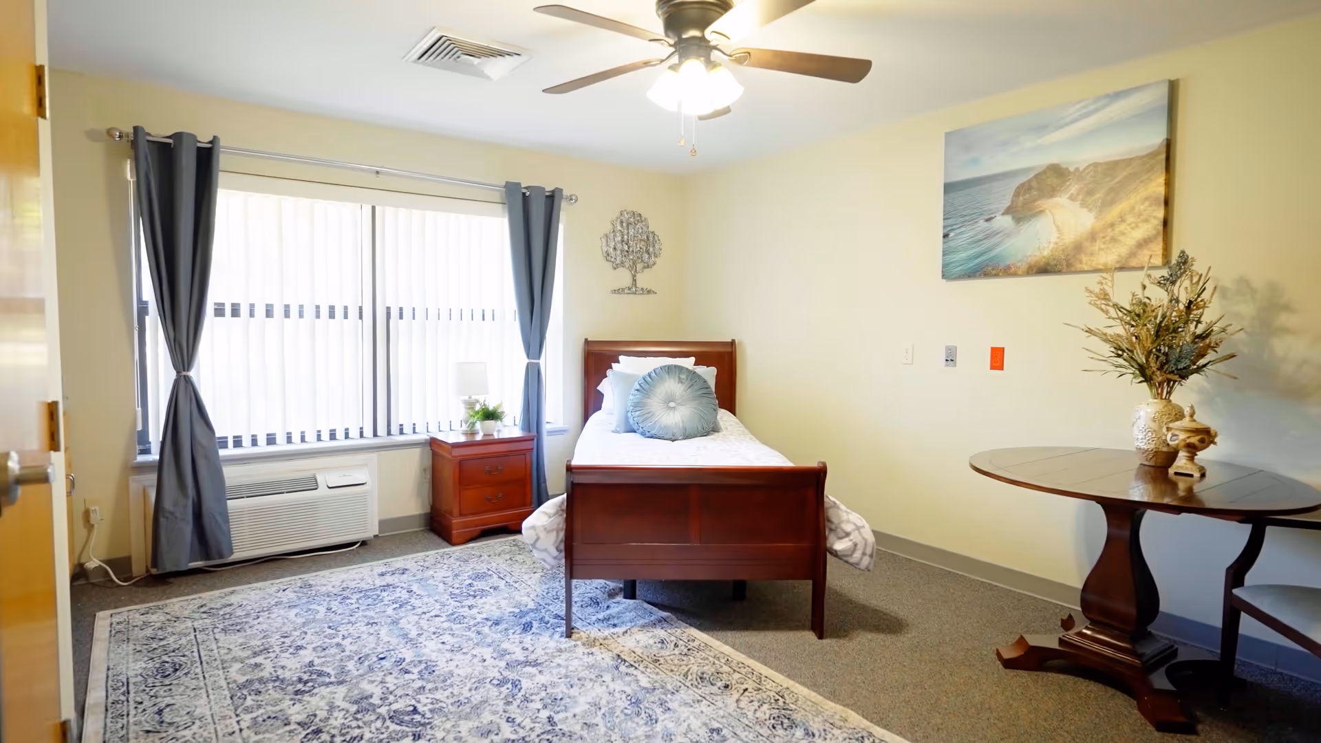 A bright and cozy bedroom in Forest Hills Assisted Living featuring a single wooden bed with white bedding and a decorative round pillow. The room has a large window with vertical blinds and blue curtains, a wooden nightstand with a lamp and plant, a ceiling fan with light, a patterned area rug, a round wooden table with a vase of flowers, and a scenic beach wall art.