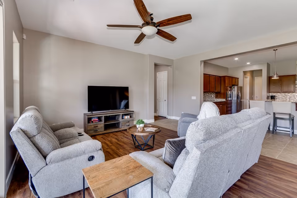 Bright open living room with gray reclining sofas, a TV on a wooden console, a ceiling fan, and a view into the kitchen.
