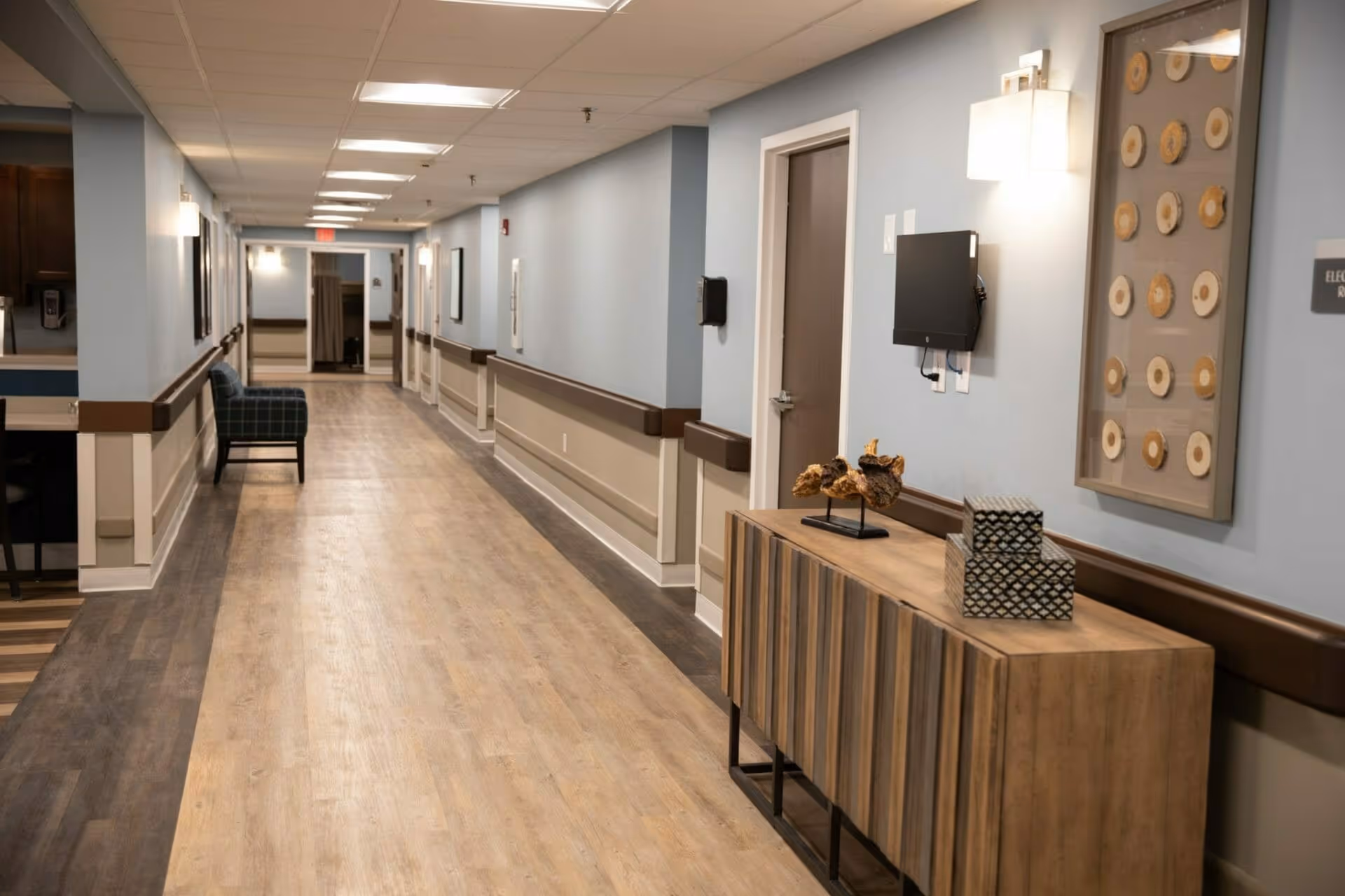 A long, well-lit hallway in a senior living facility with light blue walls and wood-patterned flooring. There is a wooden console table on the right side with decorative items, a wall-mounted monitor, and framed artwork above it. On the left side, there is a blue plaid armchair and a partial view of a sink area. The hallway has multiple doors and handrails along the walls.