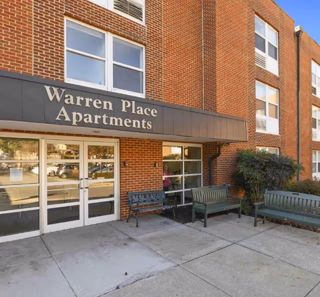 Exterior view of the entrance to Warren Place Apartments, a brick building with large windows and a black awning displaying the facility name. There are benches and some shrubbery near the entrance on a concrete sidewalk.