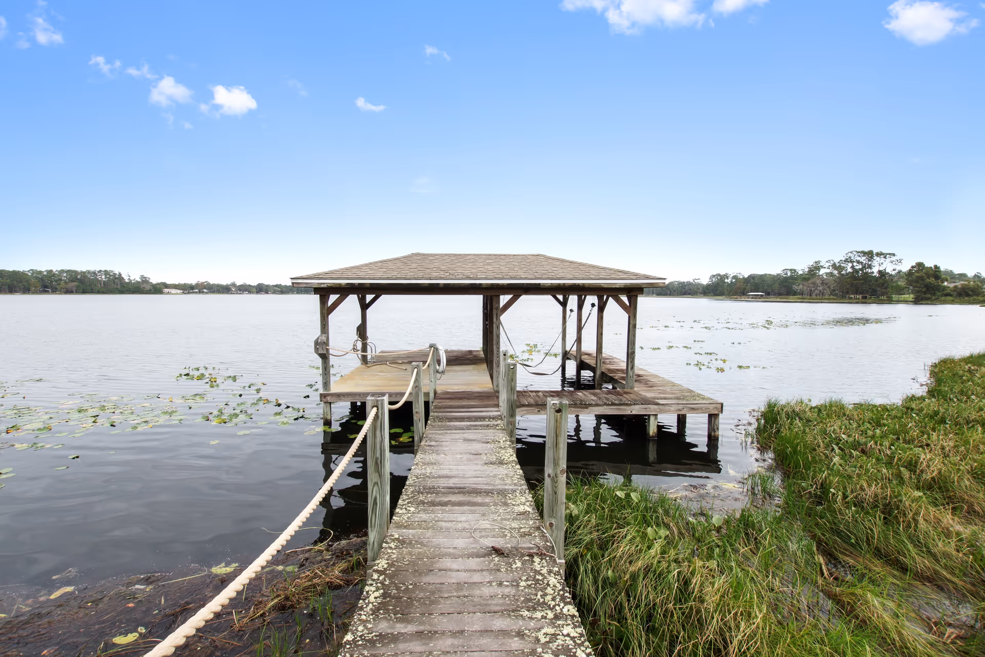 A wooden dock extending over a calm lake with a covered seating area at the end. The dock is surrounded by water with some aquatic plants and grassy shoreline visible. The sky is clear with a few clouds.