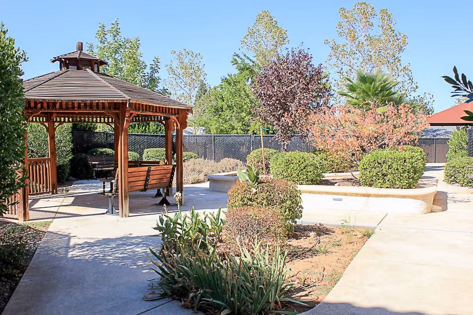 Sunlit landscaped courtyard with a wooden gazebo, benches, paved walkways, and trimmed shrubs and trees.