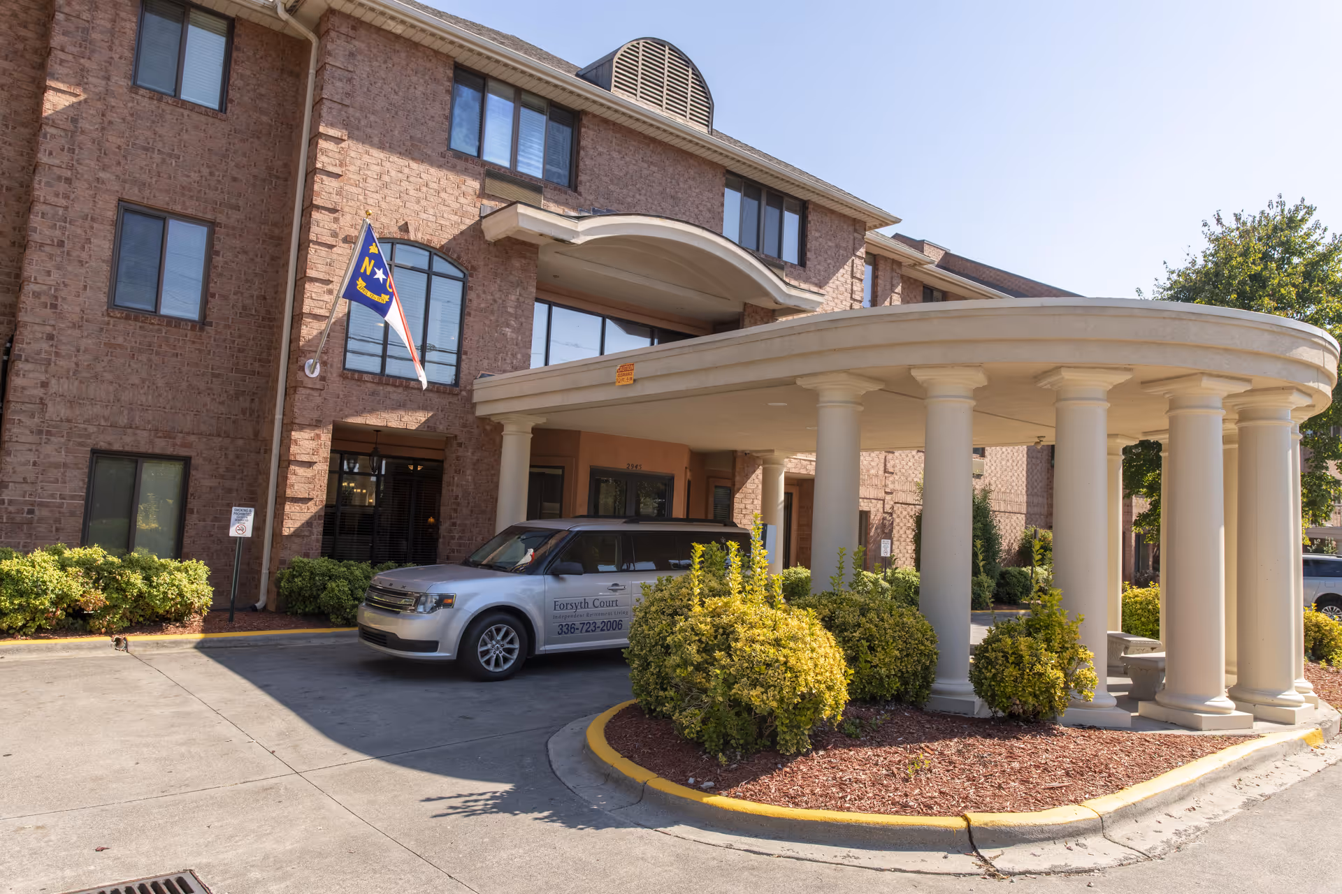 Exterior view of Holiday Forsyth Court, a brick building with multiple windows and a covered entrance supported by white columns. A silver vehicle with Forsyth Court signage is parked under the entrance canopy. There are neatly trimmed bushes and a North Carolina state flag mounted on the building.