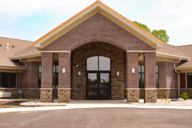 Front exterior view of CopperStone Assisted Living facility showing a brick building with a peaked roof, large glass double doors at the entrance, and stone accents at the base of the columns.
