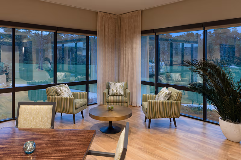 Cozy seating area with three striped armchairs around a round coffee table beside floor-to-ceiling windows overlooking a lawn.